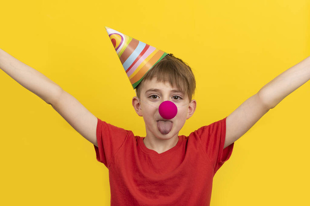 Fröhliches Kind mit roter Clownsnase und festlichem Hut auf gelbem Hintergrund. Aprilscherz - Foto, Bild