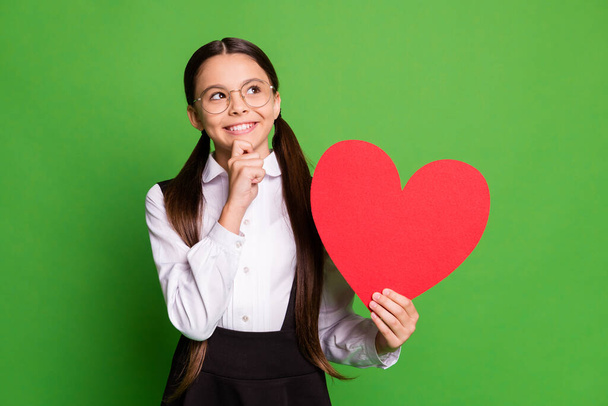 Photo of pretty cunning little schoolkid lady in front of blackboard hold show paper red heart figure look up empty space hand chin wear white shirt glasses isolated green color background - Foto, immagini