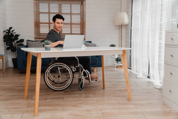 Boldog fogyatékkal élő ázsiai férfi ül tolószékben, és otthon számítógéppel dolgozik, a technológia koncepciója a fogyatékkal élők kényelmét szolgálja.. - Fotó, kép