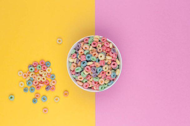 Bowl of colorful cereals top view and some cereals around.  Two colored background, yellow and pink. - Foto, Bild