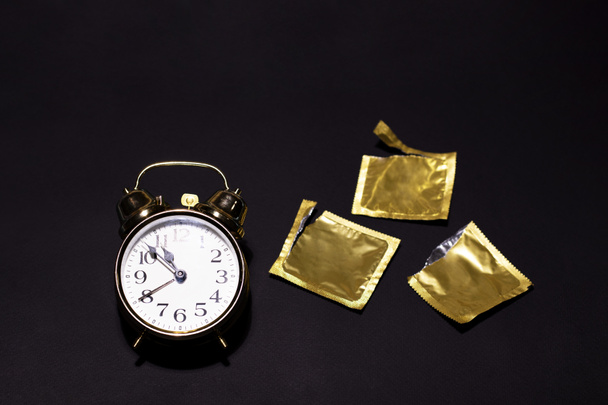 Eine goldene Uhr neben geöffneten Kondomen auf schwarzem Hintergrund - Foto, Bild