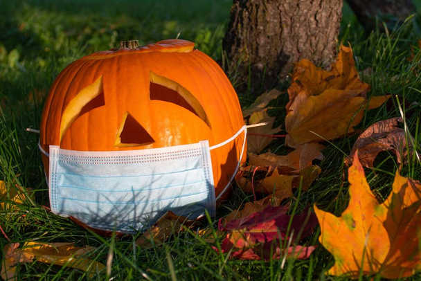 Carved Halloween pumpkin with sad eyes wearing mask on the grass with leaves, no party during Covid or Coronavirus outbreak, events and parties canceled to prevent the risk of spreading the virus - Photo, Image