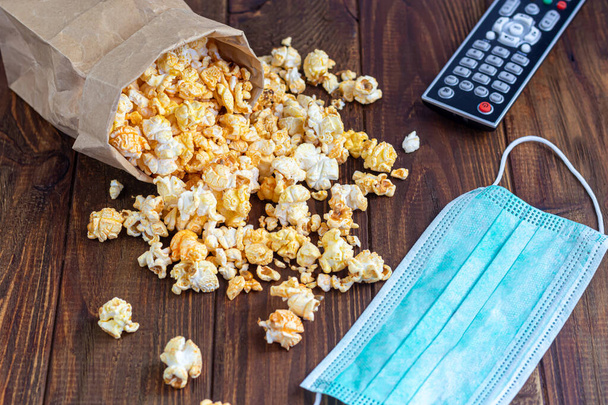 A transparent bowl of popcorn on a wooden surface. Next to a laptop and headphones. The mask protects against viruses. Copy space. - Photo, Image