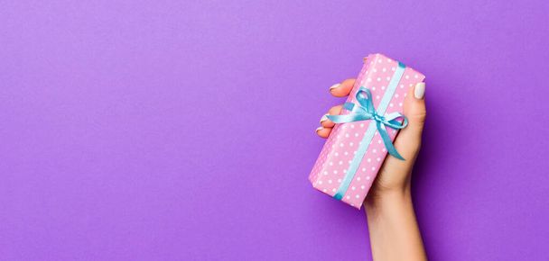 Flat lay of woman hands holding gift wrapped and decorated with bow on purple background with copy space. Christmas and holiday concept. - Photo, Image