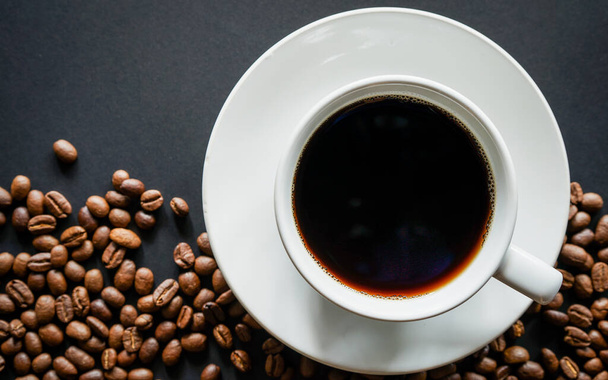 taza blanca con café caliente y granos de café marrón tostado sobre fondo oscuro, vista superior  - Foto, imagen