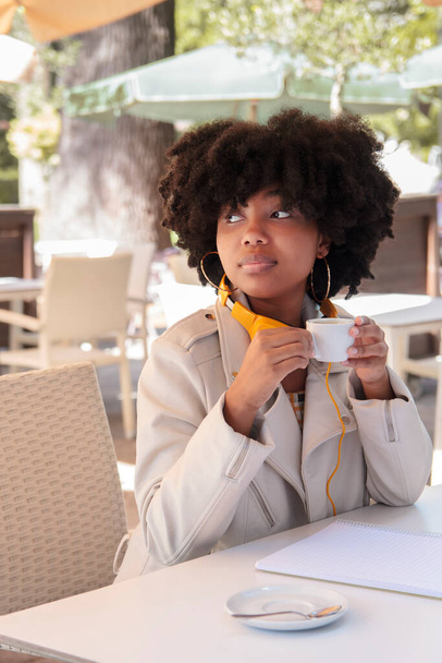 Nuori afroamerikkalainen nainen, jolla on kiharat hiukset ja takki ja kuulokkeet, juo yksin kahvia ulkoilmakahvilan terassilla aurinkoisena päivänä. College tai lukiolainen. - Valokuva, kuva
