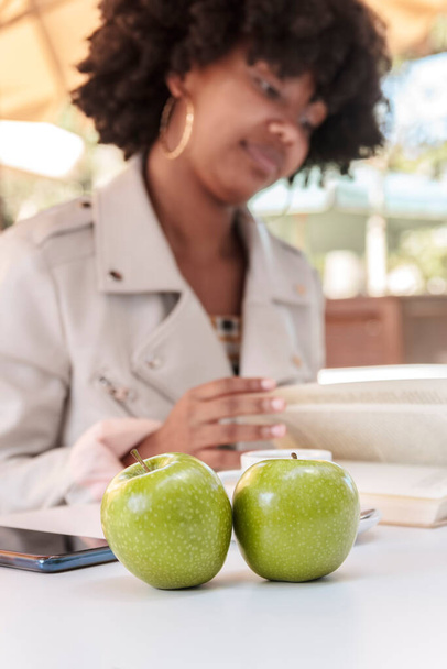 Selective focus of two green apples on a white table where a young African American woman with a jacket and curly hair reads a book in an outdoor cafeteria. College or high school student. - Photo, Image