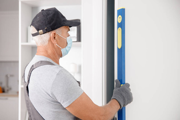 Zimmerermeister mit medizinischer Maske überprüft Tür auf Vertikalität mit Blasenwaage - Foto, Bild