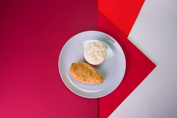 Top kilátás ízletes sült csirke filé szürke tányéron főtt fehér rizs szolgált kulináris gyűrű. Reggeli színes vörös háttérrel. Fő és oldalsó edény egy tányéron. - Fotó, kép
