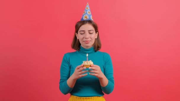 jonge vrouw in verjaardag hoed blazen kaars op taart, geïsoleerd op rode achtergrond. - Video