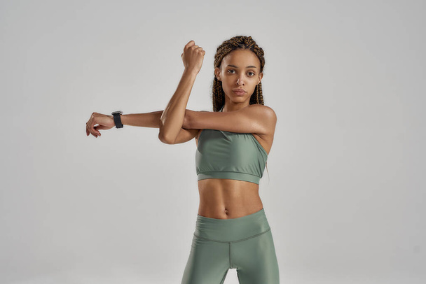 Doing sport exercises. Young mixed race fitness woman in sportswear stretching her arms before workout while standing isolated over grey background - Fotografie, Obrázek