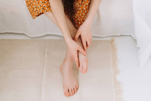 Woman sitting on the bed massages her foot, close-up. Woman with a slender body massages the leg, Young woman massaging her foot on the white bed after training or hard working day. Healthcare concept - Zdjęcie, obraz