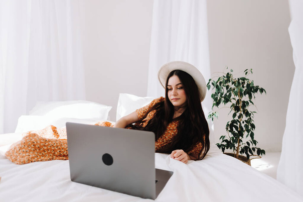 Beautiful young stylish woman in white hat lying on bed and using laptop computer in sunny morning at home. High quality photo - Zdjęcie, obraz