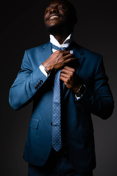 An African American man stands in a business suit and puts a tie over his head. standing on a gray background - Φωτογραφία, εικόνα