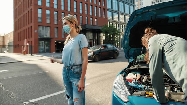 Young woman in medical mask showing hitchhiking gesture, asking for help while man examining broken down car, trying to fix it - Photo, image