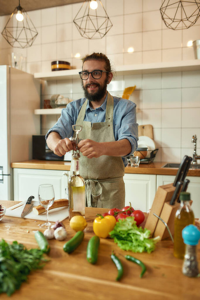 Young man, Italian cook looking cheerful while opening a bottle of white wine with a corkscrew in the kitchen - Foto, Bild