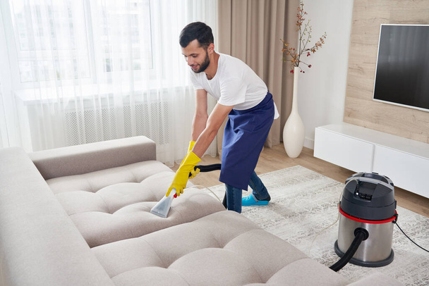 Close-up van de huishoudster met moderne wasstofzuiger en het reinigen van vuile sofa met professioneel schoonmaakmiddel. Professionele voorjaarsschoonmaak thuis concept - Foto, afbeelding