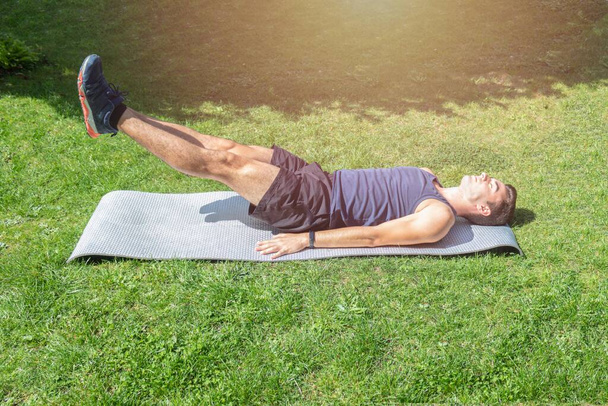 jonge man liggend op een mat doen bodybuilding oefeningen en abdominale versterking tillen zijn voeten in de tuin - Foto, afbeelding