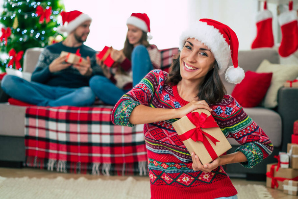 Merry Christmas and Happy New Year. Photo of a happy smiling beautiful woman in a Christmas Santa hat is showing a gift box in hands on group of friends and Christmas tree background. - Foto, afbeelding