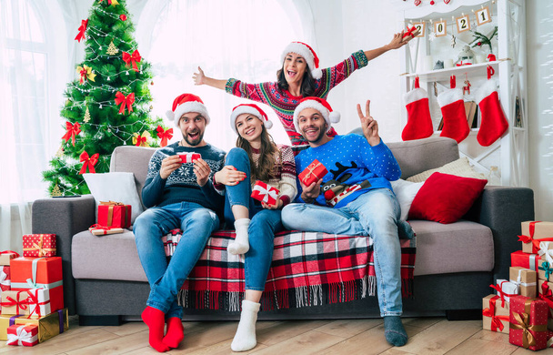 Merry Christmas and Happy New Year. The big happy excited group of young friends in Christmas santa hats with gift boxes are having fun together while sitting on the couch. - Foto, Imagen