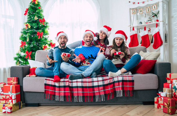 Merry Christmas and Happy New Year. The big happy excited group of young friends in Christmas santa hats with gift boxes are having fun together while sitting on the couch. - Foto, Imagem