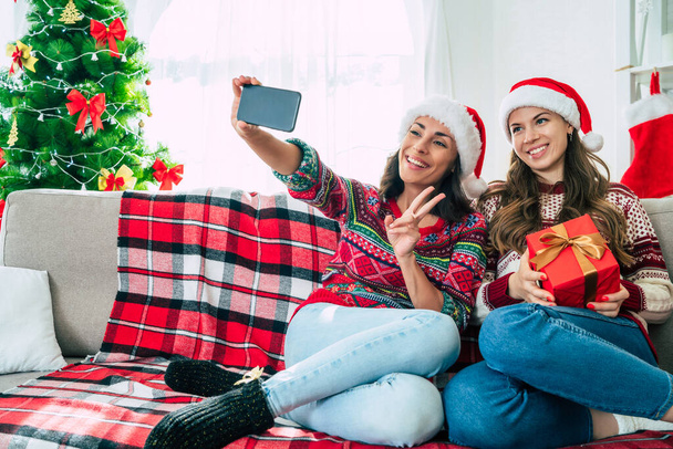 Happy New Year and Merry Christmas. Two beautiful excited young sisters or friends in Santa hats are making selfie photo on smartphone on the couch with Christmas gift boxes. - Photo, Image