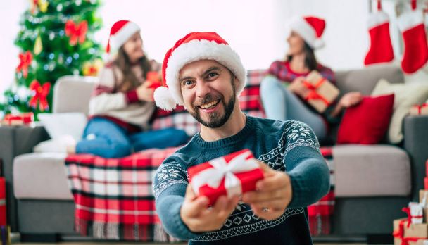Merry Christmas and Happy New Year. Young happy bearded man in a Christmas Santa hat is showing a gift box in hands on group of friends and Christmas tree background. - Valokuva, kuva
