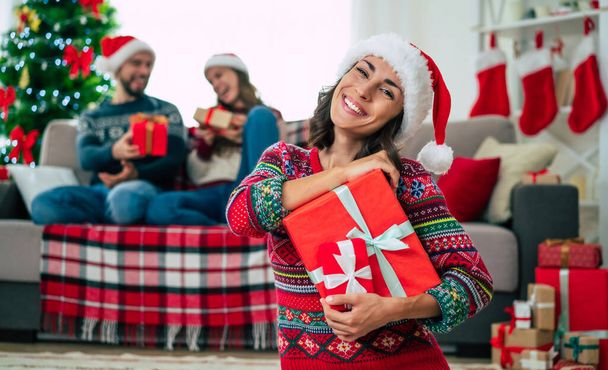 Merry Christmas and Happy New Year. Photo of a happy smiling beautiful woman in a Christmas Santa hat is showing a gift box in hands on group of friends and Christmas tree background. - Photo, image