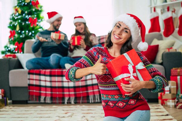 Merry Christmas and Happy New Year. Photo of a happy smiling beautiful woman in a Christmas Santa hat is showing a gift box in hands on group of friends and Christmas tree background. - 写真・画像