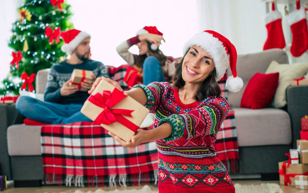 Merry Christmas and Happy New Year. Photo of a happy smiling beautiful woman in a Christmas Santa hat is showing a gift box in hands on group of friends and Christmas tree background. - Fotó, kép