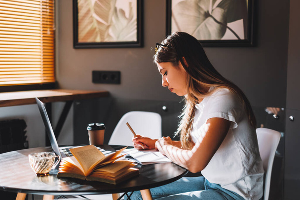 Focused caucasian young woman college student studying with books laptop distantly preparing for test exam writing essay doing homework at home, distantly education concept. - Foto, Imagen
