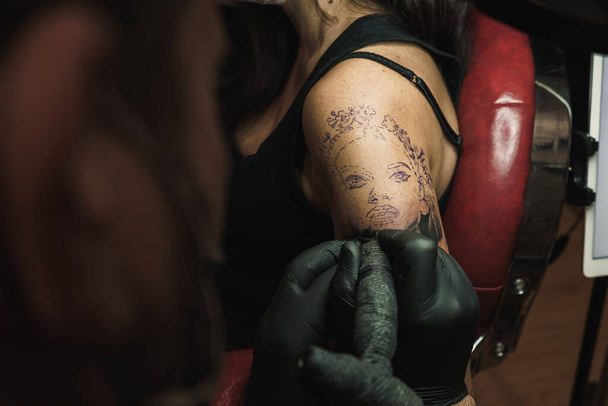 Bras de femme tatoué avec une machine protégée contre le COVID. Mode de vie. Tatoueur portant des gants en latex noir. Le tatouage est un visage de femme. - Photo, image
