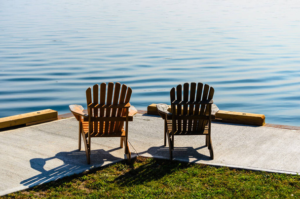 İki Adirondack sandalyesi St. Lawrence Nehri 'ndeki beton iskelede suyun kenarına bakıyor. - Fotoğraf, Görsel