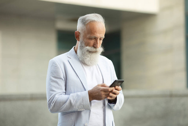 Barbudo sênior sorridente empresário em pé ao ar livre e lendo em seu telefone celular. - Foto, Imagem