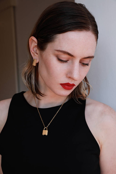 Vertical close-up of beautiful young woman with red lips and golden jewellery posing over white wall background. - Zdjęcie, obraz