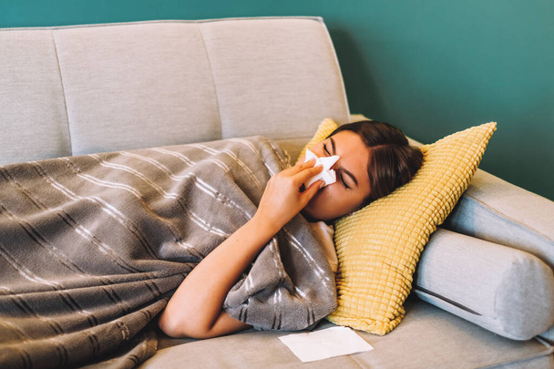 Sick young woman blowing her nose using facial tissues while lying on the sofa.  - Foto, Bild