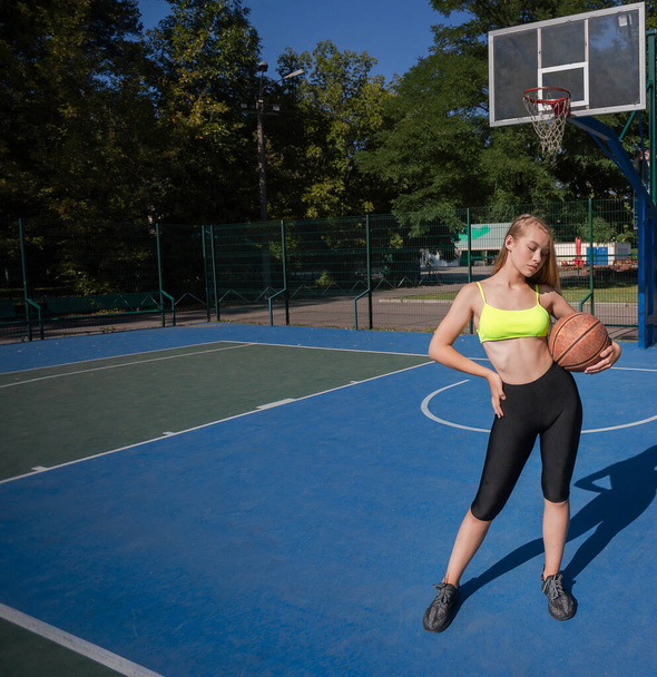 Fit adolescente sosteniendo y posando con el baloncesto en la cancha en el día soleado. Longitud completa - Foto, imagen
