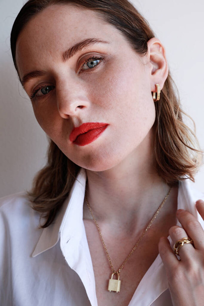 Vertical close-up portrait of beautiful woman with red lips and golden jewellery posing over white wall background. - Fotó, kép