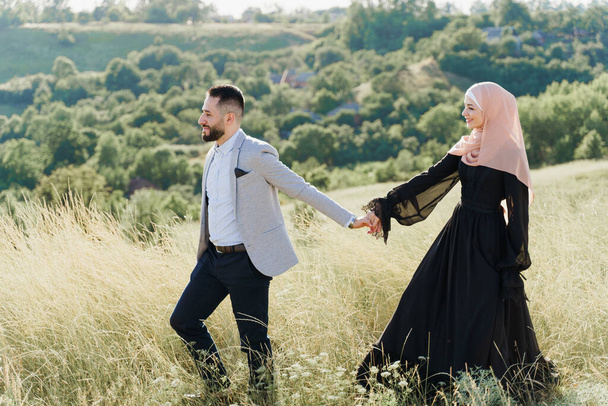 混合カップルのイスラム教徒の愛の物語。男と女の笑顔と緑の丘の上を歩く。ヒジャーブで身に着けていた女は男に見える。オンラインのための広告日付代理店 - 写真・画像
