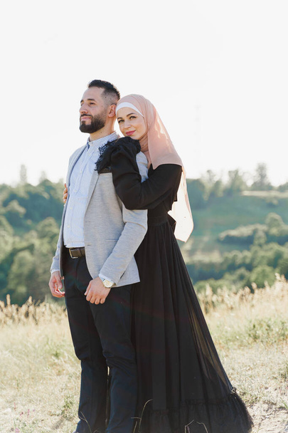 Historia de amor musulmana de cerca. Pareja mixta sonríe y abraza en las verdes colinas. La mujer vestida con hijab mira a su hombre. Anuncio de agencia de citas on-line - Foto, Imagen