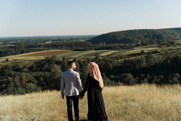 Muslim love story. Mixed couple smiles and hugs on the green hills . Woman weared in hijab looks to her man. Advert for on-line dating agency - 写真・画像