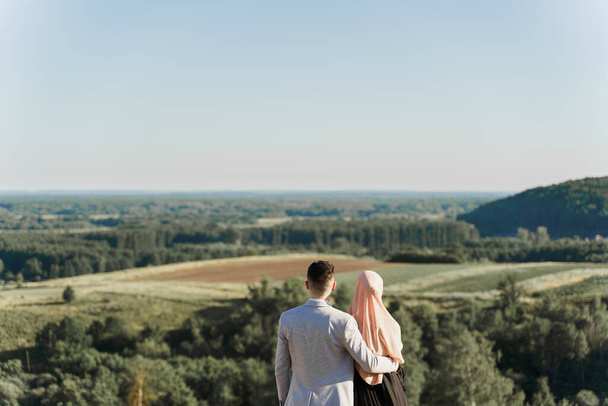 Muslimische Liebesgeschichte. Gemischtes Paar lächelt und umarmt sich auf den grünen Hügeln. Frau im Hidschab blickt zu ihrem Mann. Werbung für Online-Partnervermittlung - Foto, Bild