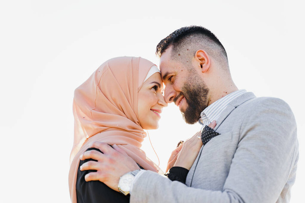 Historia de amor musulmana de cerca con la luz del sol. Pareja mixta sonríe y abraza al atardecer. La mujer vestida con hijab mira a su hombre. Anuncio de agencia de citas on-line - Foto, imagen