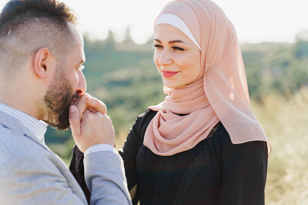 Storia d'amore musulmana primo piano. Coppie miste di sorrisi e abbracci sulle verdi colline. La donna indossata in hijab guarda al suo uomo. Pubblicità per on-line incontri agenzia - Foto, immagini
