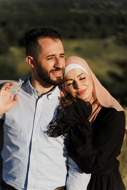 Muslim love story close-up. Mixed couple smiles and hugs on the green hills . Woman weared in hijab looks to her man. Advert for on-line dating agency - Photo, Image