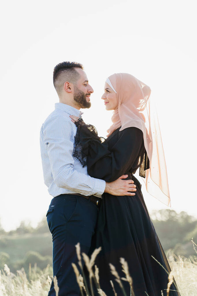 Moslim liefdesverhaal met zonlicht. Gemengde paar glimlachen en knuffels bij zonsondergang. Een vrouw in hijab kijkt naar haar man. Advertentie voor online dating bureau - Foto, afbeelding