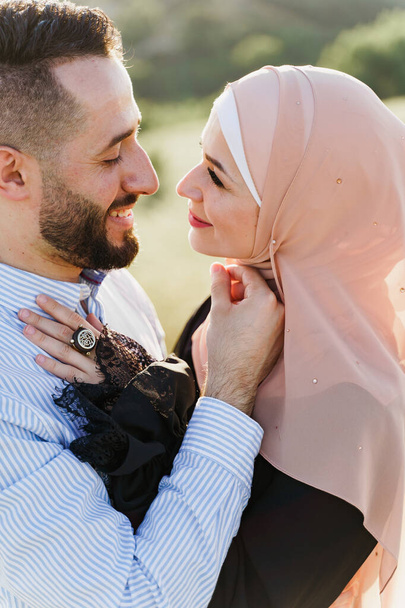 Muslim love story close-up. Mixed couple smiles and hugs on the green hills . Woman weared in hijab looks to her man. Advert for on-line dating agency - Foto, imagen