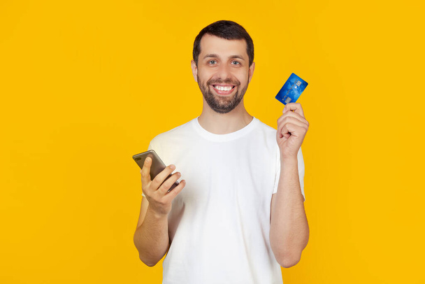 Young man with a beard in a white t-shirt holding a credit card. by credit card to pay online using a smartphone. Stands on isolated yellow background. - Zdjęcie, obraz