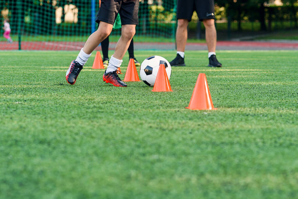 Close up boy in sportswear trains football on soccer field and learns to circle the ball between training orange cones. - Foto, Imagen