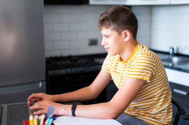 Cute young teenager in white shirt sitting behind desk in kitchen next to laptop and study. Serious boy in earphones makes homework, listening lesson. Home, distance education, self study by kids. - Фото, зображення